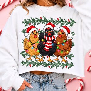 Puede incluir: Una sudadera blanca de manga larga con un estampado gr&aacute;fico de tres pollos con gorros de Pap&aacute; Noel rojos y blancos. Los pollos est&aacute;n envueltos en luces de Navidad de colores y el pollo central lleva una bufanda a rayas rojas y blancas.