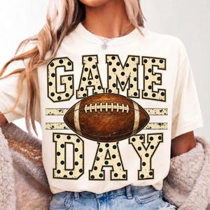 Op de afbeelding: Een cr&egrave;mekleurig T-shirt met korte mouwen en de tekst GAME DAY in grote blokletters met een zwart stippenpatroon. In het midden tussen de woorden staat een afbeelding van een bruine American football met witte veters.
