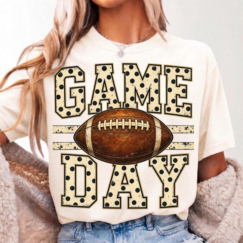Op de afbeelding: Een cr&egrave;mekleurig T-shirt met korte mouwen en de tekst GAME DAY in grote blokletters met een zwart stippenpatroon. In het midden tussen de woorden staat een afbeelding van een bruine American football met witte veters.