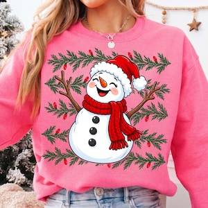 Puede incluir: Una sudadera de color rosa brillante con cuello redondo que presenta un gr&aacute;fico central de un mu&ntilde;eco de nieve sonriente con bufanda roja y gorro de Pap&aacute; Noel rojo. El mu&ntilde;eco tiene brazos de rama y tres botones negros sobre ramas de pino.
