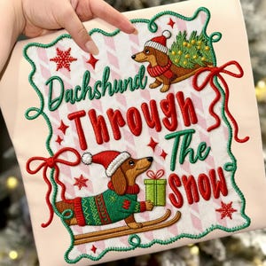 Może przedstawiać: Haftowana poduszka z napisem "Dachshund Through The Snow". Projekt przedstawia jamniki w czapkach i swetrach Świętego Mikołaja, niosące choinkę i prezent. Poduszka ma różowo-białe wzorzyste tło z czerwonymi i zielonymi akcentami.
