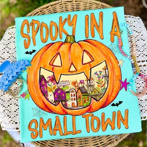 May include: A light blue canvas print featuring a large orange jack-o'-lantern with a carved face. Inside the pumpkin is a colorful illustration of a small town with houses, ghosts, and flowers. The text reads SPOOKY IN A SMALL TOWN.