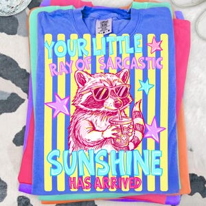 May include: A stack of colorful t-shirts. The top shirt is blue with a graphic of a raccoon wearing sunglasses and drinking from a cup. Text on the shirt reads "YOUR LITTLE RAY OF SARCASTIC SUNSHINE HAS ARRIVED."