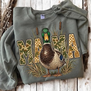 May include: An olive green crewneck sweatshirt featuring a graphic of a mallard duck standing among tall grasses and cattails. The word MAMA is printed in large block letters with a camouflage and checkered pattern overlaying the design.