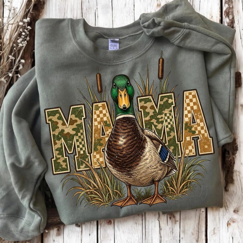 May include: An olive green crewneck sweatshirt featuring a graphic of a mallard duck standing among tall grasses and cattails. The word MAMA is printed in large block letters with a camouflage and checkered pattern overlaying the design.