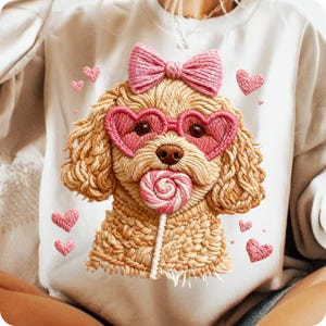 May include: A cream-colored sweatshirt featuring an embroidered dog wearing heart-shaped sunglasses and a bow. The dog is holding a lollipop and surrounded by pink heart accents. The design has a textured, stitched appearance.