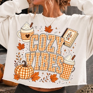 Puede incluir: Una sudadera de manga larga color crema con un estampado gr&aacute;fico que incluye el texto COZY VIBES en cuadros vichy naranjas y blancos. El dise&ntilde;o presenta una calabaza, dos tazas con nata montada, un libro y hojas de oto&ntilde;o en tonos naranja y marr&oacute;n.