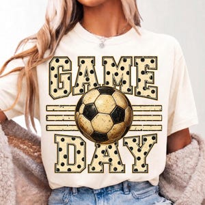 May include: A cream-colored short-sleeve t-shirt featuring a graphic print with the text GAME DAY in large block letters filled with black polka dots. A soccer ball with a distressed texture is positioned between the words GAME and DAY.
