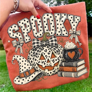 May include: An orange t-shirt featuring the word SPOOKY in large white polka-dot letters. The graphic includes a leopard-print pumpkin with a black and white plaid bow, a polka-dot witch hat, a black cauldron with a heart, and a stack of three books.