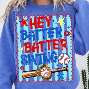 Może przedstawiać: Kr&oacute;lewsko-niebieska bluza z okrągłym dekoltem i grafiką w jasnoniebieskie pionowe paski z czerwoną falistą obw&oacute;dką. Napis HEY BATTER BATTER SWING jest w kolorze czerwonym i niebieskim, z ilustracjami piłki baseballowej oraz rękawicy.