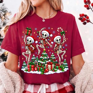 Op de afbeelding: Een bordeauxrood t-shirt met een grafische print van drie skeletten met kerstmutsen en sjaals, dansend rond versierde kerstbomen met cadeautjes en een zuurstok, inclusief kleurrijke kerstverlichting en sneeuwvlokken.