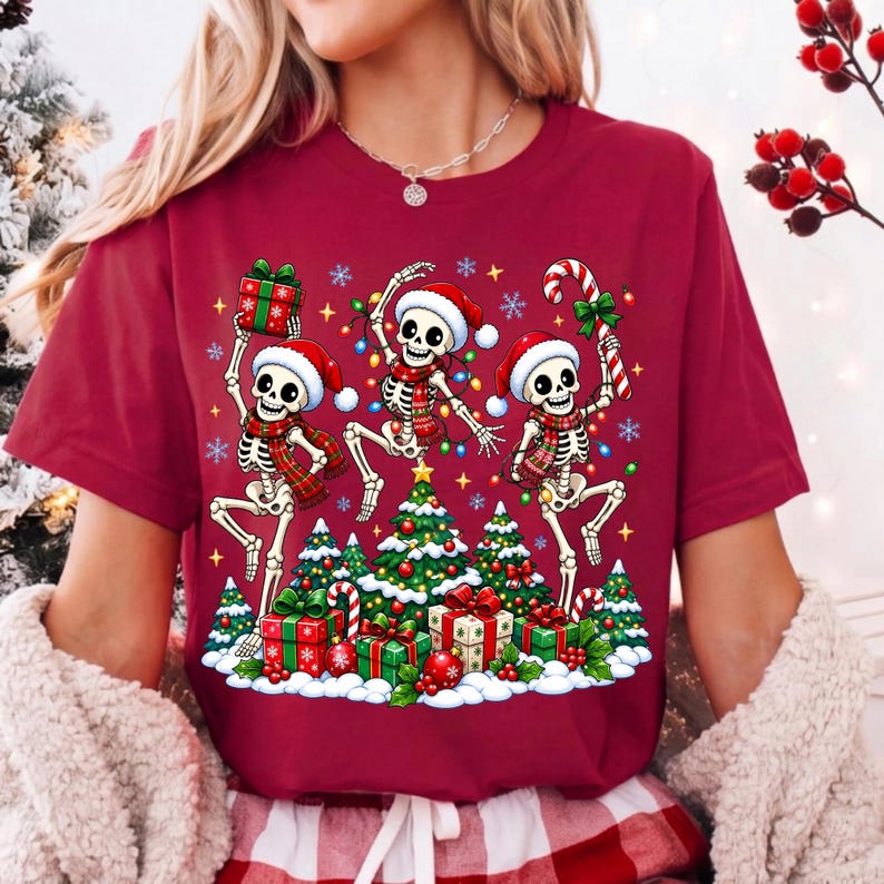 Op de afbeelding: Een bordeauxrood t-shirt met een grafische print van drie skeletten met kerstmutsen en sjaals, dansend rond versierde kerstbomen met cadeautjes en een zuurstok, inclusief kleurrijke kerstverlichting en sneeuwvlokken.
