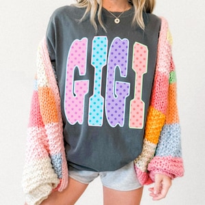 May include: Dark gray sweatshirt with the word "GIGI" in large, colorful, polka-dotted letters. The letters are outlined in white. A multi-colored, patchwork cardigan is draped over the shoulders.