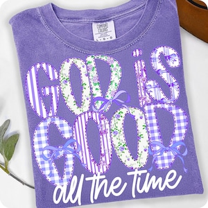 K&ouml;nnte beinhalten: Ein lilafarbenes T-Shirt mit dem Schriftzug "GOD IS GOOD all the time". Die Buchstaben sind mit Streifen, floralen Mustern und Vichy-Karo gestaltet und mit blauen Schleifen verziert. Das Shirt hat einen Rundhalsausschnitt und kurze &Auml;rmel.