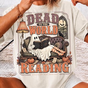 May include: A cream-colored t-shirt featuring a graphic of a ghost sitting on a purple sofa reading a book. The text reads "DEAD TO THE WORLD" and "READING" with "please do not resurrect" below. The design includes a lamp, pumpkins, and a cat.