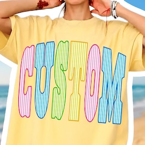 May include: A pale yellow short-sleeve t-shirt featuring the word CUSTOM printed in large, bold, arched letters. Each letter has a vertical pinstripe pattern in alternating colors of pink, blue, green, and yellow, creating a vibrant graphic design.