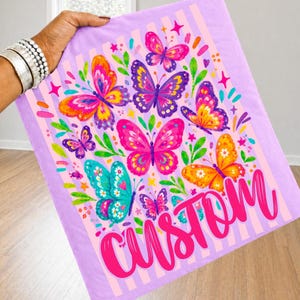 May include: A light purple fabric featuring a pattern of colourful butterflies in shades of pink, orange, yellow, and teal, accented with green leaves and small stars. The word CUSTOM is printed in large, bold, bright pink script at the bottom.