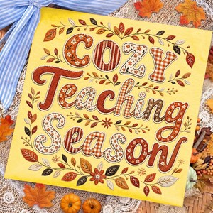 May include: A square yellow sign featuring the text Cozy Teaching Season in a decorative font. The letters are filled with patterns of orange, brown, and white plaid, stripes, and floral motifs, surrounded by a border of autumn leaves and vines.