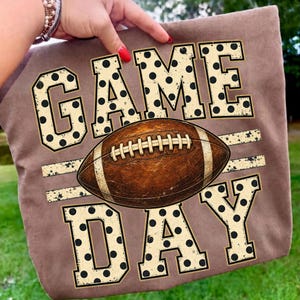 Op de afbeelding: Een bruin t-shirt met de tekst GAME DAY in grote, cr&egrave;mekleurige blokletters met zwarte stippen. Een bruine American football met witte veters is horizontaal geplaatst tussen de woorden GAME en DAY.