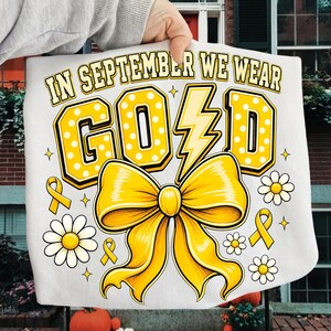 May include: A white t-shirt featuring the text IN SEPTEMBER WE WEAR GOLD in yellow block letters with white polka dots. The letter L is replaced by a yellow lightning bolt. A large yellow bow, small yellow ribbons, and white daisies are printed.