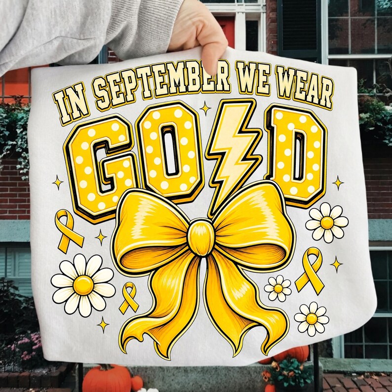 May include: A white t-shirt featuring the text IN SEPTEMBER WE WEAR GOLD in yellow block letters with white polka dots. The letter L is replaced by a yellow lightning bolt. A large yellow bow, small yellow ribbons, and white daisies are printed.