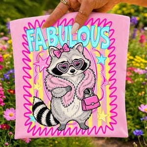 K&ouml;nnte beinhalten: Ein hellrosa Stoffartikel mit einem Cartoon-Waschb&auml;ren, der eine herzf&ouml;rmige Brille, eine rosa Schleife und eine rosa Pelzstola tr&auml;gt und eine rosa Handtasche h&auml;lt. Das Wort FABULOUS steht in gro&szlig;en blauen Buchstaben oben, umgeben von Sternen.