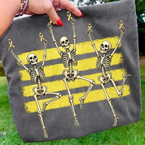 May include: A charcoal grey tote bag featuring a graphic print of three skeletons in dynamic poses. Each skeleton holds a yellow awareness ribbon above its head. The design includes three horizontal yellow brushstroke stripes behind the skeletons.
