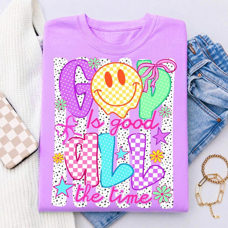 May include: A light purple t-shirt featuring a graphic design with the text "God is good all the time". The design includes a yellow checkered smiley face, colorful checkered letters, pink bows, small stars, and flowers over a black dotted background.