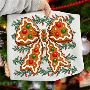 Op de afbeelding: Een wit t-shirt met een grote afbeelding van een peperkoekstrik versierd met wit glazuur, rode en groene snoepjes, omringd door een patroon van groene dennentakken en kleine rode kerstlichtjes.