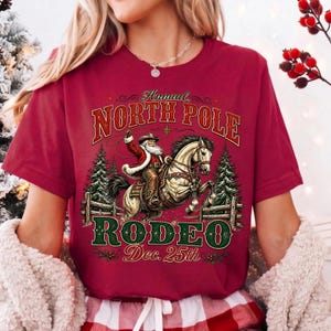 May include: A maroon short-sleeve t-shirt featuring a graphic of Santa Claus riding a horse with a lasso. The text reads Annual NORTH POLE RODEO Dec. 25th. The design includes two pine trees, a wooden fence, and decorative holly accents.