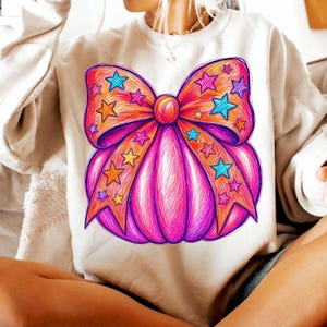 May include: A beige crewneck sweatshirt featuring a large, vibrant graphic of a pumpkin adorned with a prominent bow. The design is rendered in a scribbled, sketch-like style with bright pink, orange, and blue colors, accented by numerous small stars.