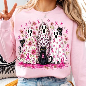 May include: A light pink sweatshirt featuring a graphic print of three ghosts and three black cats. The ghosts are decorated with pink floral, leopard, and polka dot patterns. Pink breast cancer awareness ribbons are scattered throughout the design.