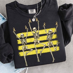 May include: A black crewneck sweatshirt featuring a graphic of three dancing skeletons. Each skeleton holds a small gold awareness ribbon above its head. Behind the skeletons are three horizontal yellow brushstroke stripes. The inner label reads Comfort Colors.