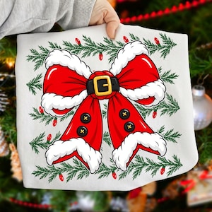 Puede incluir: Una camiseta blanca con un gr&aacute;fico de un gran lazo rojo con borde de piel sint&eacute;tica blanca, un cintur&oacute;n negro con hebilla dorada y cuatro botones negros. El dise&ntilde;o se sit&uacute;a sobre un fondo de ramas de pino verdes y luces navide&ntilde;as rojas.