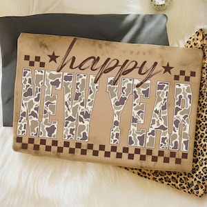 May include: A rectangular pillow with the words "happy NEW YEAR" in brown lettering. The letters "NEW YEAR" are filled with a camouflage pattern in shades of brown, gray, and beige. The pillow has a brown checkered border.