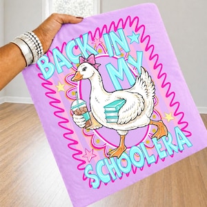 May include: A light purple blanket featuring a cartoon goose with a pink bow, holding a drink and books. The blanket has the text "BACK IN MY SCHOOLERA" in blue and pink, with stars and a wavy border.