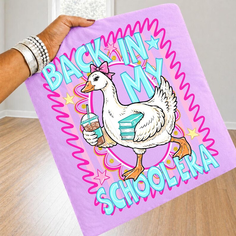 May include: A light purple blanket featuring a cartoon goose with a pink bow, holding a drink and books. The blanket has the text "BACK IN MY SCHOOLERA" in blue and pink, with stars and a wavy border.