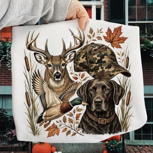 May include: A white sweatshirt featuring a graphic print of a deer, a mallard duck in flight, a camouflage baseball cap, and a black Labrador retriever wearing a collar, surrounded by cattails and autumn maple leaves.