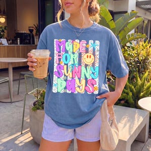 May include: A blue short-sleeve t-shirt featuring a graphic print with the text "MADE FOR SUNNY DAYS" in colorful, patterned bubble letters. The design includes a yellow smiley face, small flowers, and stars against a background of vertical stripes.