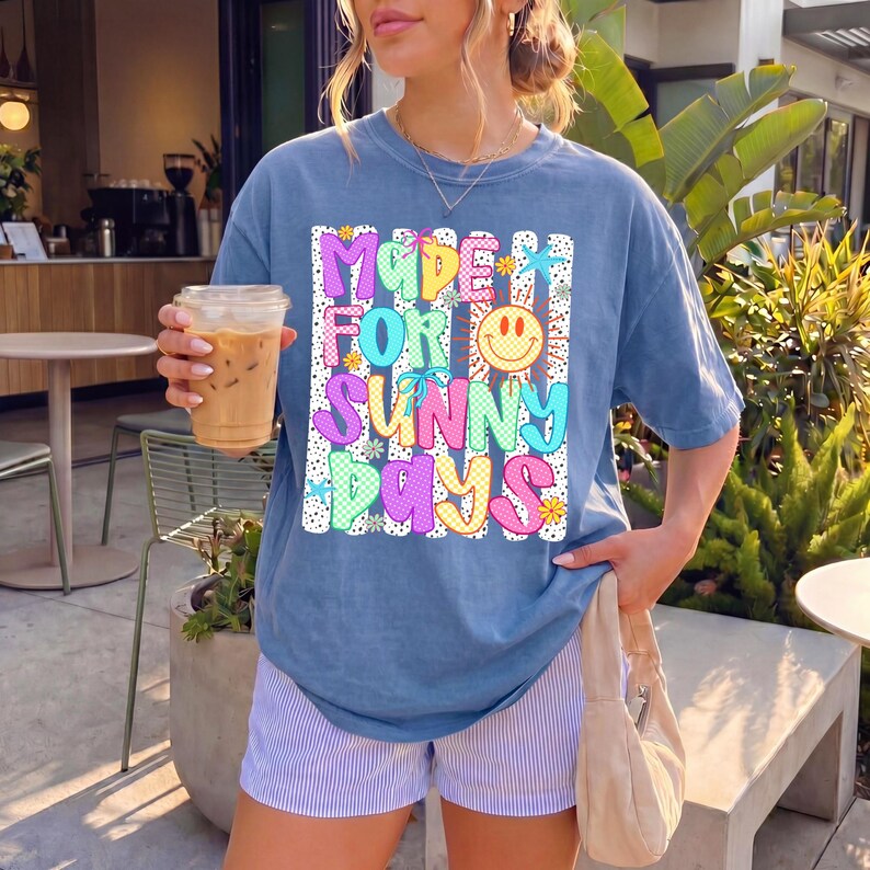 May include: A blue short-sleeve t-shirt featuring a graphic print with the text "MADE FOR SUNNY DAYS" in colorful, patterned bubble letters. The design includes a yellow smiley face, small flowers, and stars against a background of vertical stripes.