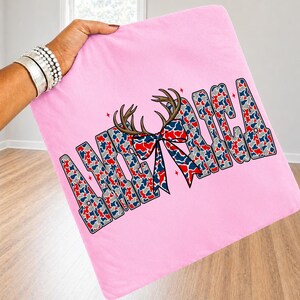 May include: A light pink fabric square featuring the word AMERICA in large block letters filled with a red, white, and blue camouflage pattern. A brown deer antler graphic with a matching red, white, and blue patterned bow sits in the center.