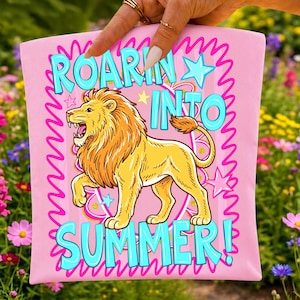 Puede incluir: Un art&iacute;culo de tela rosa claro con una ilustraci&oacute;n central de un le&oacute;n dorado con una melena completa. El texto "ROARIN INTO SUMMER!" est&aacute; impreso en letras grandes de color azul claro. El dise&ntilde;o incluye bordes ondulados rosas y estrellas azules.