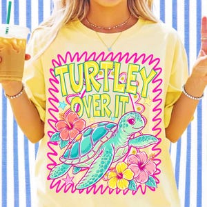 K&ouml;nnte beinhalten: Ein blassgelbes T-Shirt mit einem Grafikdruck einer Meeresschildkr&ouml;te, umgeben von rosa und gelben Hibiskusbl&uuml;ten. Der Text "TURTLEY OVER IT" ist in gro&szlig;en gelben Buchstaben &uuml;ber der Schildkr&ouml;te gedruckt, alles in einem leuchtend rosa Rahmen.