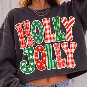 May include: A charcoal gray cropped sweatshirt featuring the text HOLLY JOLLY in large, multi-patterned letters. The letters include red bows, green polka dots, red and white gingham, green Christmas trees, and red and white candy cane stripes.