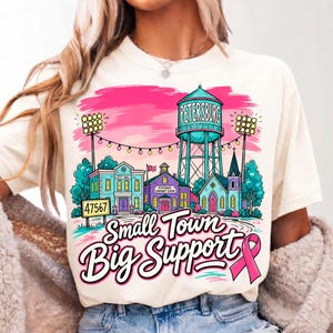 May include: A cream-colored t-shirt featuring a colorful graphic of a small town with a water tower labeled Petersburg, a city hall, a school, and a church. The design includes string lights, stadium floodlights, and the text Small Town Big Support.