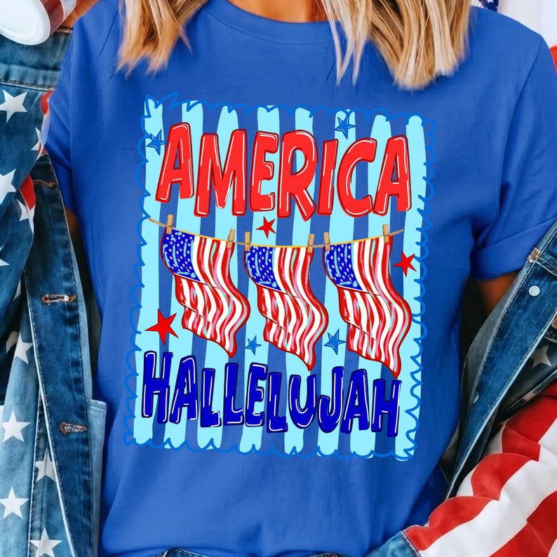 Puede incluir: Una camiseta azul real con un estampado gr&aacute;fico de tres banderas estadounidenses colgadas en un tendedero. El texto AMERICA aparece en rojo sobre las banderas y HALLELUJAH en azul debajo, sobre un fondo de rayas de color azul claro.