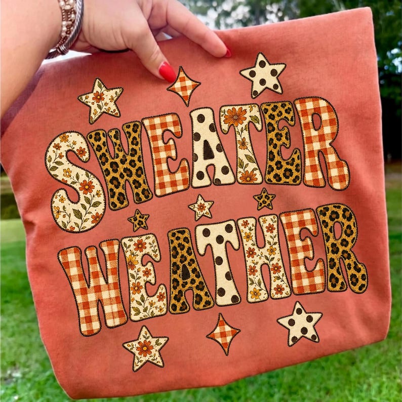 May include: A dusty rose fabric item featuring the text "SWEATER WEATHER" in large, bold letters. The letters are filled with various patterns including orange gingham, leopard print, floral motifs, and polka dots, accented by small star shapes.