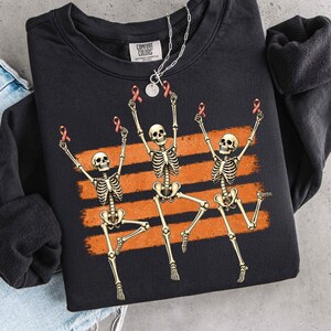 May include: A black crewneck sweatshirt featuring a graphic of three dancing skeletons. Each skeleton holds a small red awareness ribbon above its head. The design is set against three horizontal orange brushstroke stripes. The inner label reads Comfort Colors.