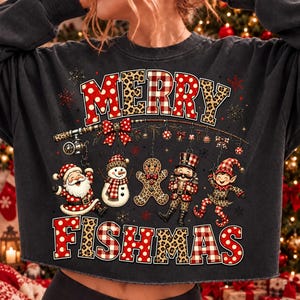 May include: A dark grey sweatshirt featuring the text MERRY FISHMAS in red and white plaid and leopard print. The design includes a fishing rod with ornaments of Santa, a snowman, a gingerbread man, a nutcracker, and an elf hanging from it.