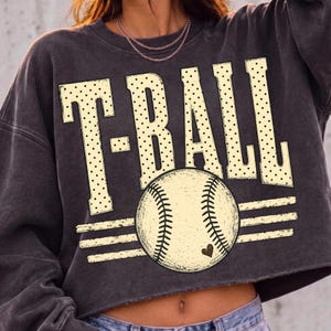 Pode incluir: Uma camisola curta cinzento-carv&atilde;o com o texto T-BALL em letras grandes de cor creme com um padr&atilde;o de pontos pretos. Abaixo do texto, encontra-se um gr&aacute;fico de uma bola de basebol com costuras pretas e um pequeno cora&ccedil;&atilde;o preto.